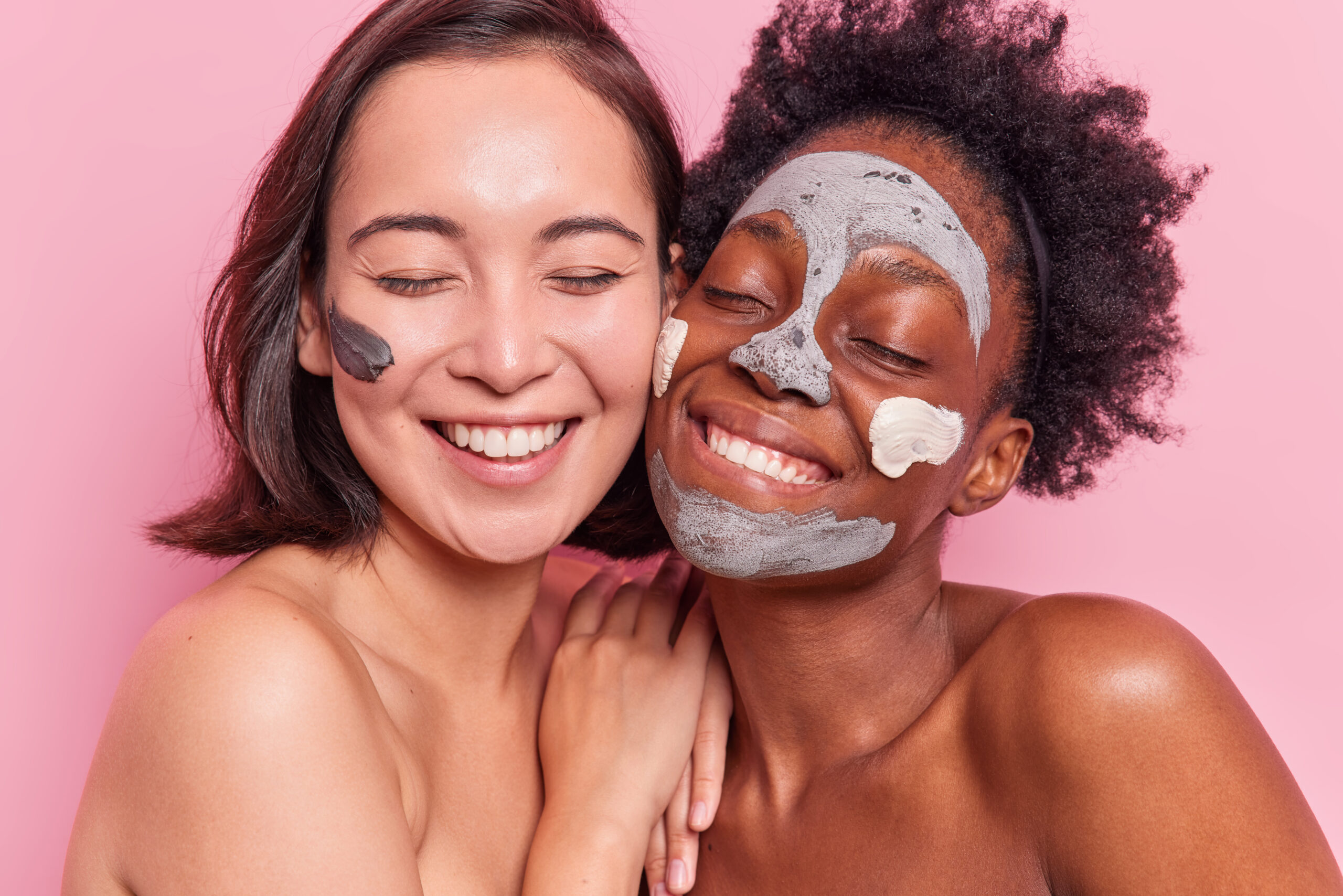Portrait of happy sincere diverse young women smile toothily apply clay mask on face undergo beauty procedures stand shirtless next to each other with naked body isolated over rosy background.